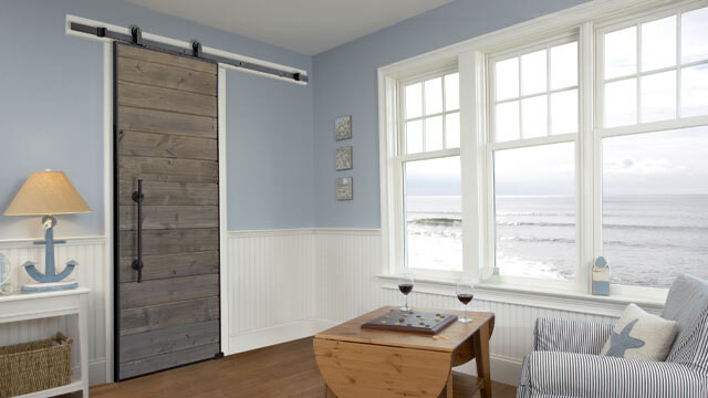 A rustic sliding barn door in a weathered driftwood finish adds character to a coastal-themed room with light blue walls, white wainscoting, and ocean-view windows. The black hardware and vertical plank design of the door contrast beautifully with the soft, beachy décor. Nautical accents, a cozy sitting area, and a scenic ocean backdrop complete the relaxed, seaside ambiance.