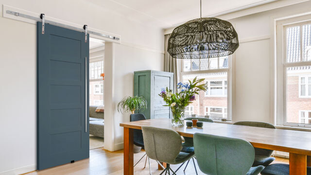 A contemporary dining space features a sleek MDF Shaker 5-panel barn door in a bold deep blue finish, mounted with a white sliding track. The room is filled with natural light from large windows, showcasing a blend of modern and organic elements, including a wood dining table, green upholstered chairs, and a woven pendant light. The door adds a stylish and functional design element, seamlessly connecting adjoining spaces.