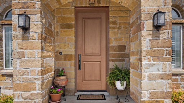 A beautiful front entry featuring a warm-toned woodgrain fiberglass door set in a stone archway. The door is complemented by a matte black handle set and surrounded by neutral, earth-toned stonework. Flanking the entry are decorative potted plants and lantern-style wall sconces, enhancing the inviting, nature-inspired aesthetic of the home.